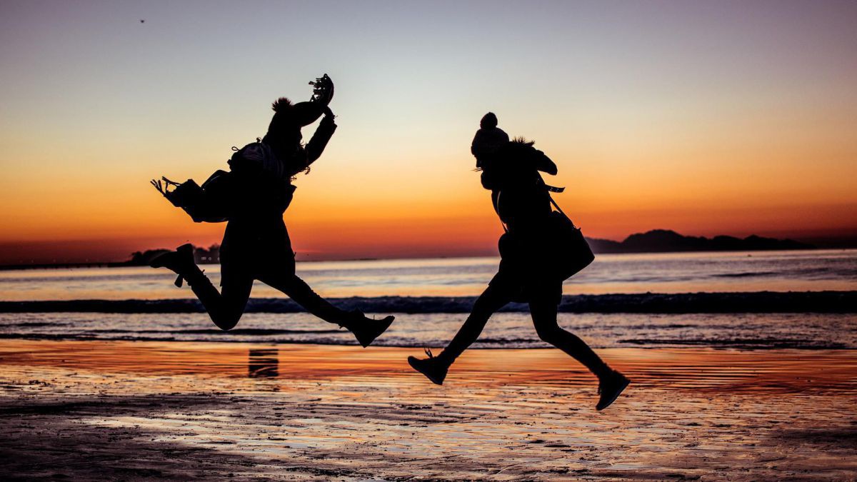 Zwei Menschen springen an einem Strand in den Sonnenuntergang. Sie sind nur als Silhouetten vor dem bunten Farbenspiel erkennbar.