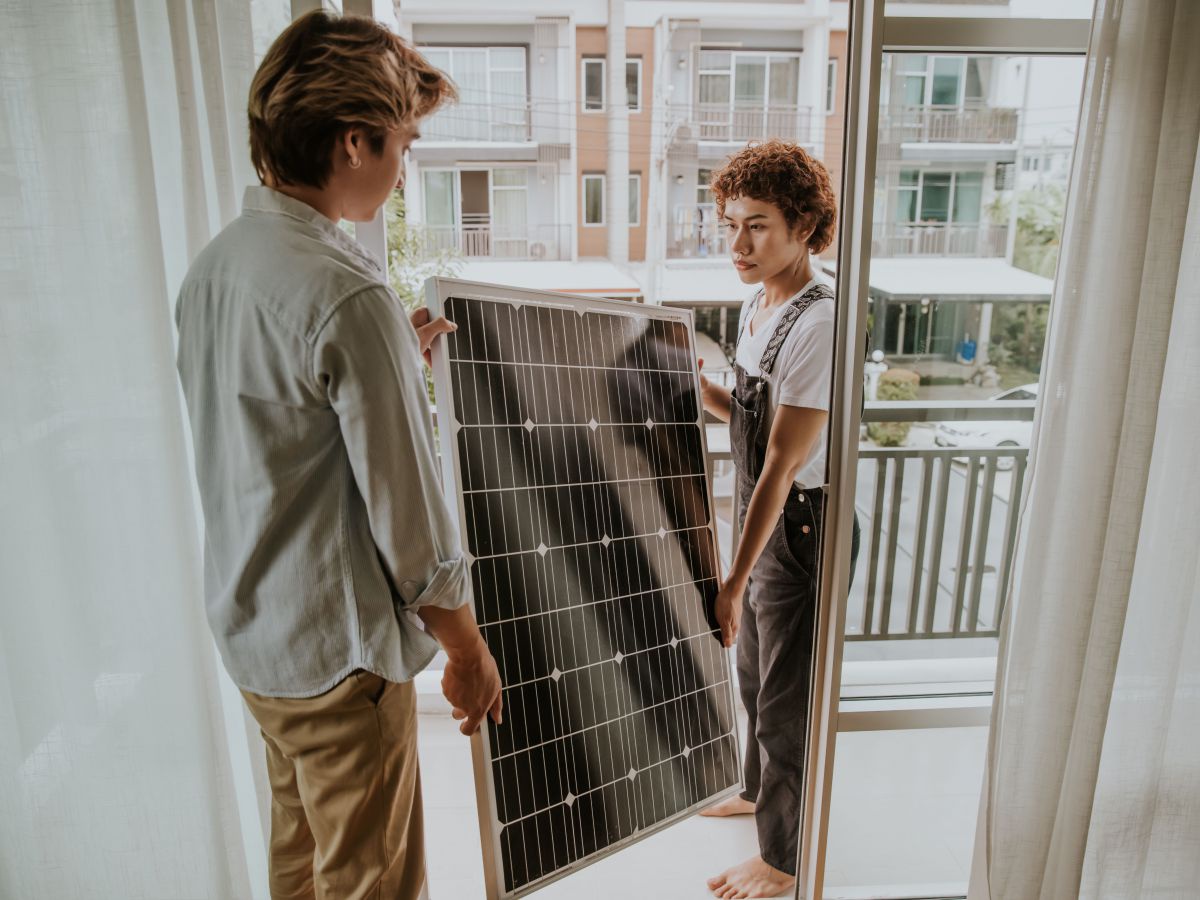Zwei junge Männer tragen gemeinsam ein Solarpanel durch die Türe auf ihren Balkon.