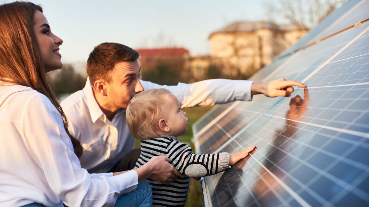 Eine junge Familie mit Kleinkind inspiziert gemeinsam eine Photovoltaikanlage, das Kind berührt das Panel, der Vater zeigt mit dem Finger erklärend auf die Zellen.