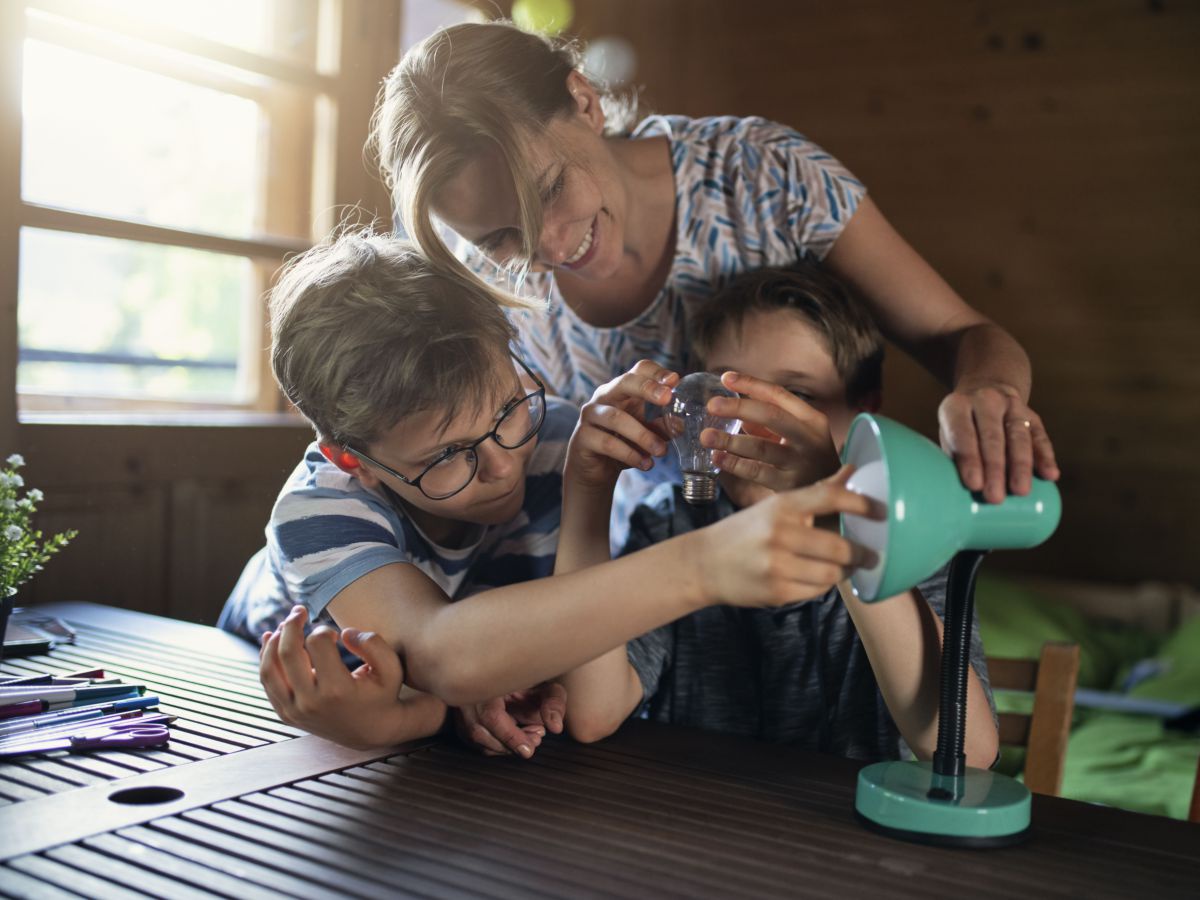 Eine Mutter mit zwei Kindern im Schulalter in einem Wohnraum, sie tauschen gerade gemeinsam die Glühbirne einer Schreibtischlampe aus.