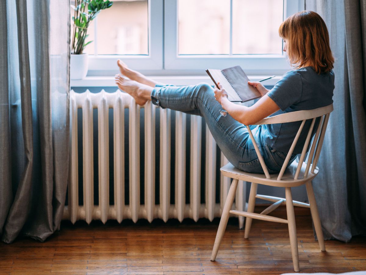 Eine Frau sitzt behaglich in einem Holzsessel am Fenster und liest in einem Notizbuch, die Beine hat sie auf dem Heizkörper abgelegt.