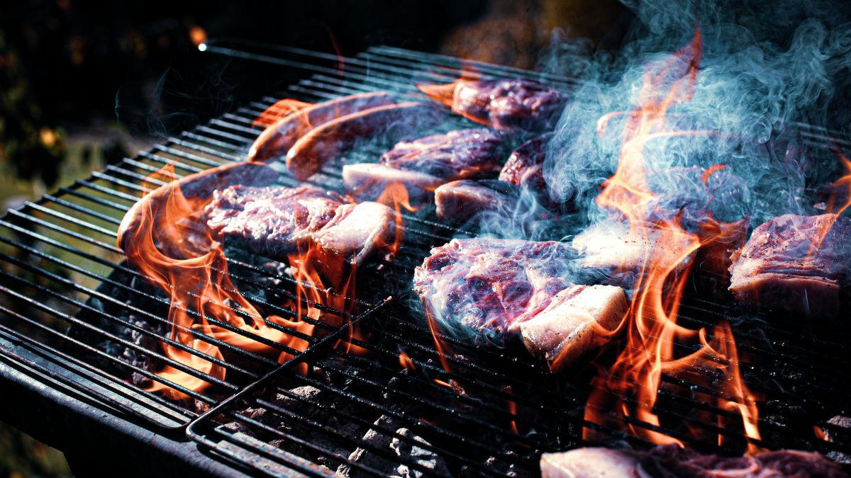 Steaks und Würste auf einem Grillrost, durch den die Flammen züngeln und blauer Rauch aufsteigt.