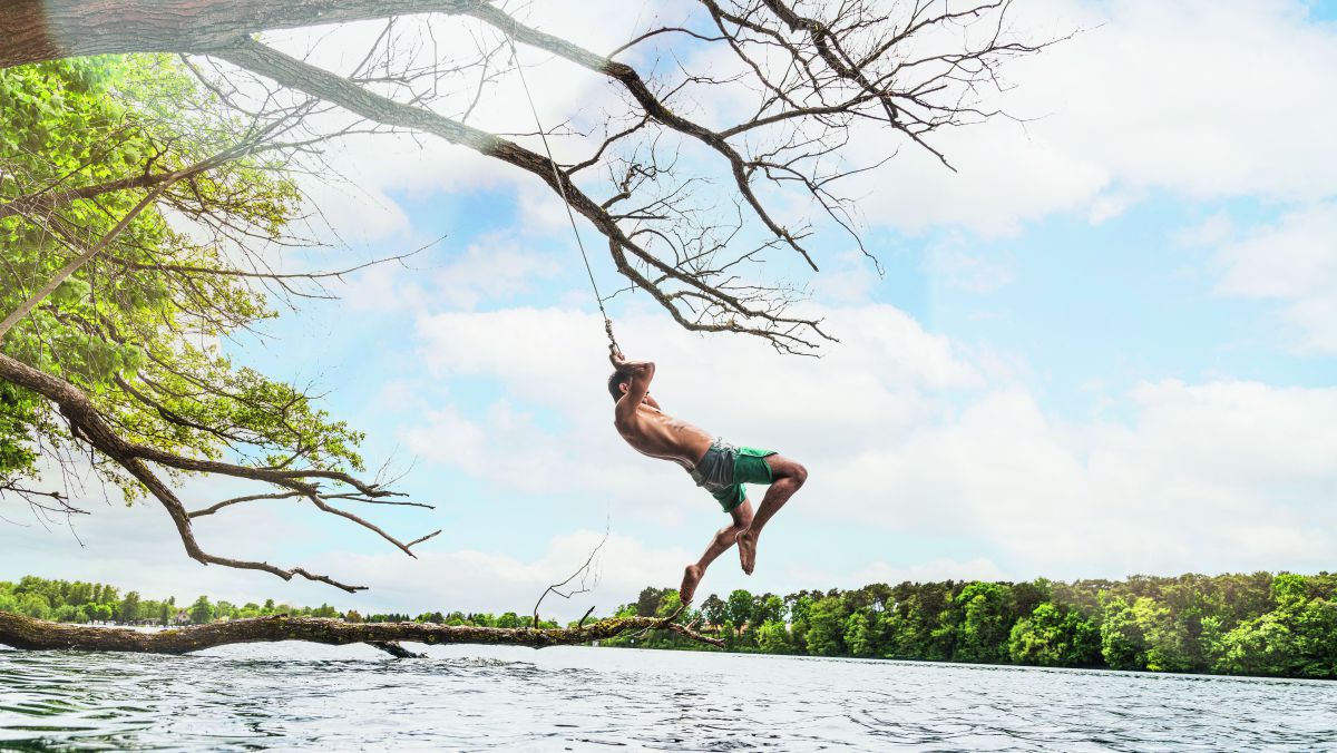 Ein junger Mann in Badehose schwingt sich mit einem Seil von einem Baum über die Wasserfläche eines Naturgewässers.