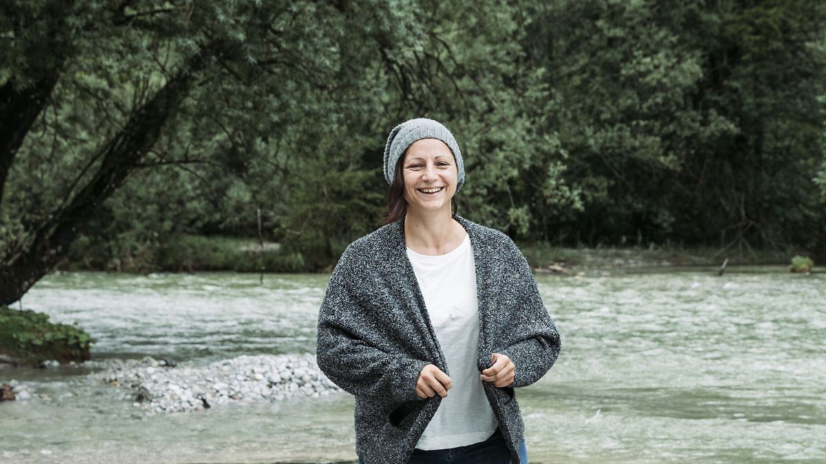 Die Autorin Maria Kapeller mit Wollweste und Haube, im Hintergrund ein von Bäumen gesäumter türkisgrüner Fluss.