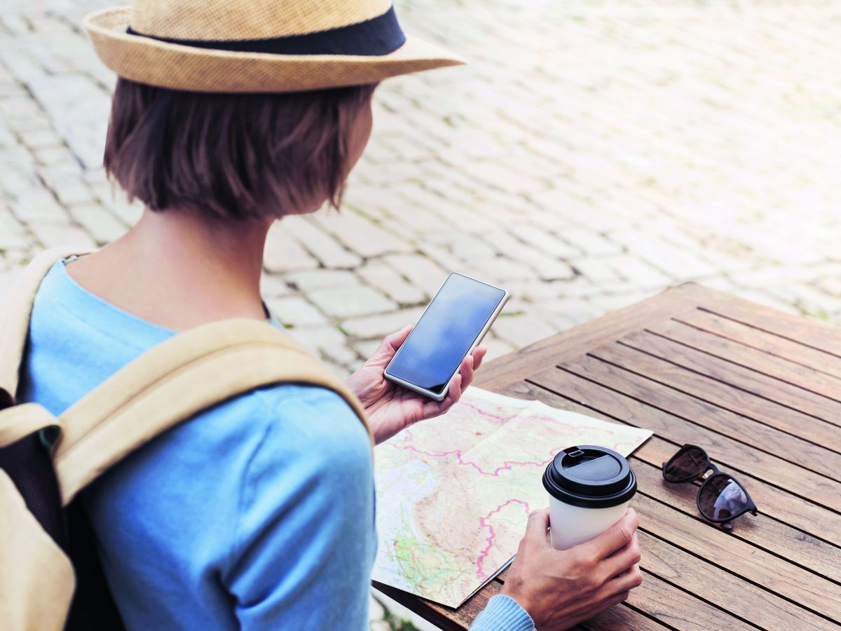 Eine Urlauberin mit Sonnenhut und Rucksack sitzt mit einem Becher Coffee-to-go an einem Bistrotisch, Landkarte und Sonnenbrille griffbereit, in der linken Hand hält sie ihr Smartphone.