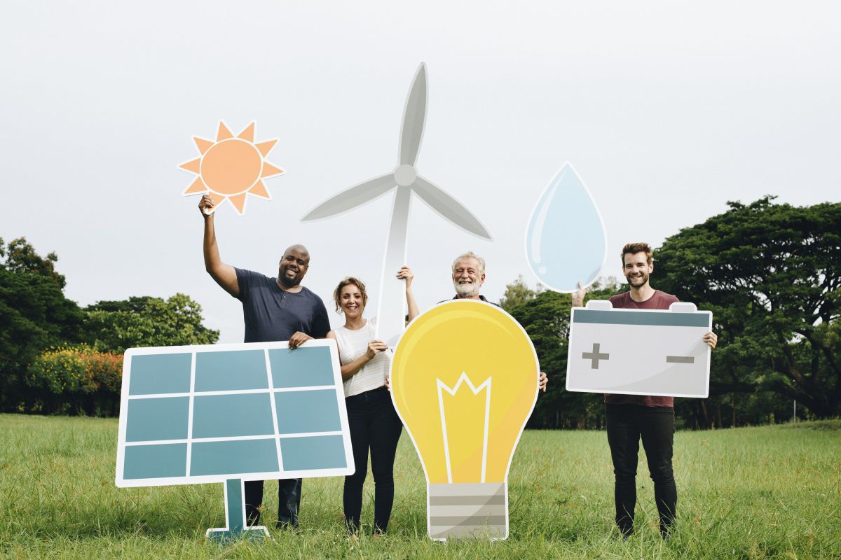 Vier Menschen stehen auf einer grünen Wiese und halten symbolhafte Papierschilder einer Sonne, einer Glühbirne, eines Windrads, einer Photovoltaikanlage, eines Wassertropfens und einer Batterie hoch.