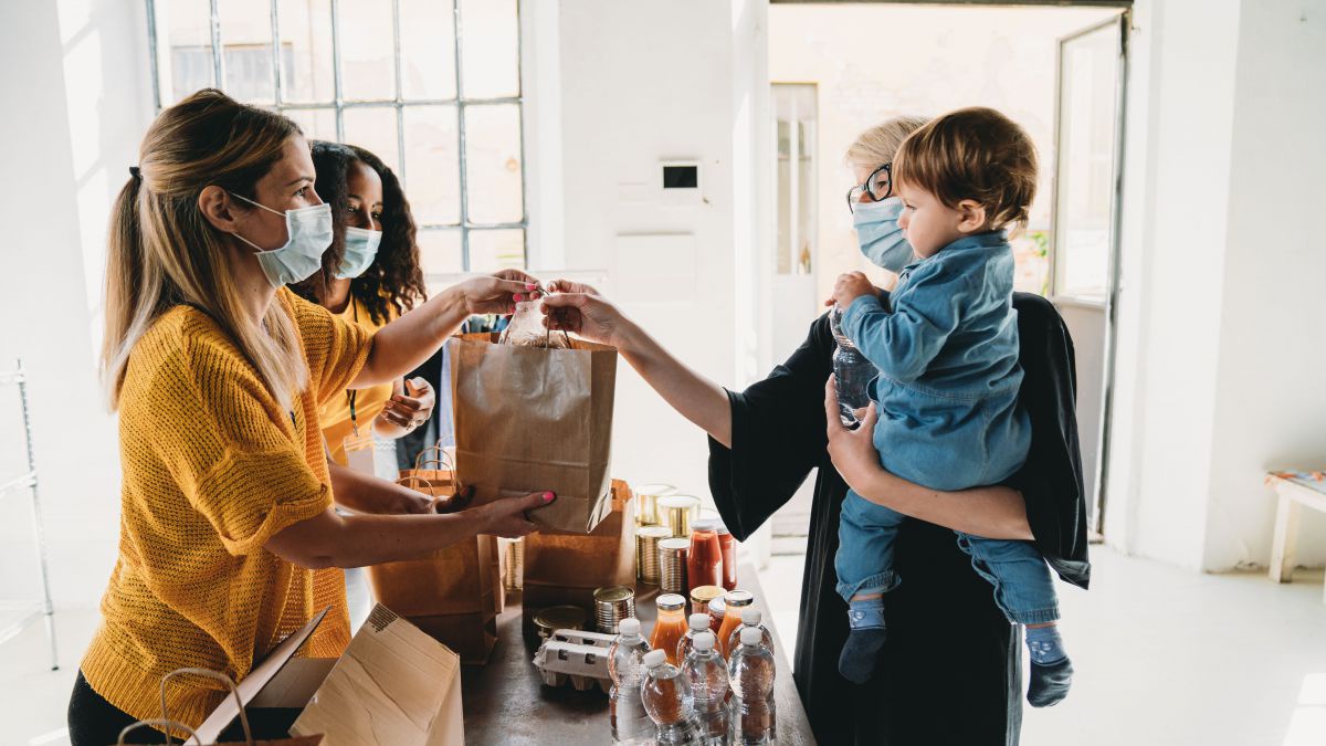 Bei einer Essensausgabe übergibt eine junge Frau eine Papiertüte Essen an eine Frau mit einem Kind auf dem Arm. Alle tragen Mund-Nasen-Schutz.