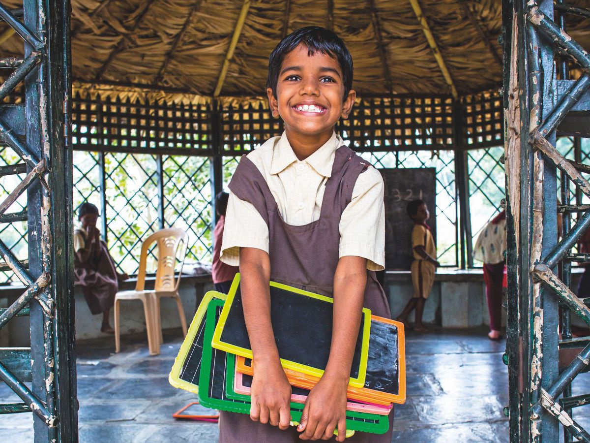 Ein Kind steht barfuß am einfang einer runden Hütte. Es hat Schulkreidetafeln in den Händen und lacht.