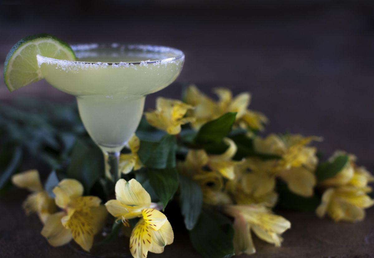 Ein blasgelber Drink in einer Cocktailschale steht inmitten von gelben Blüten. Das Glas hat einen Salzrand, an ihm steckt eine Limettenscheibe.