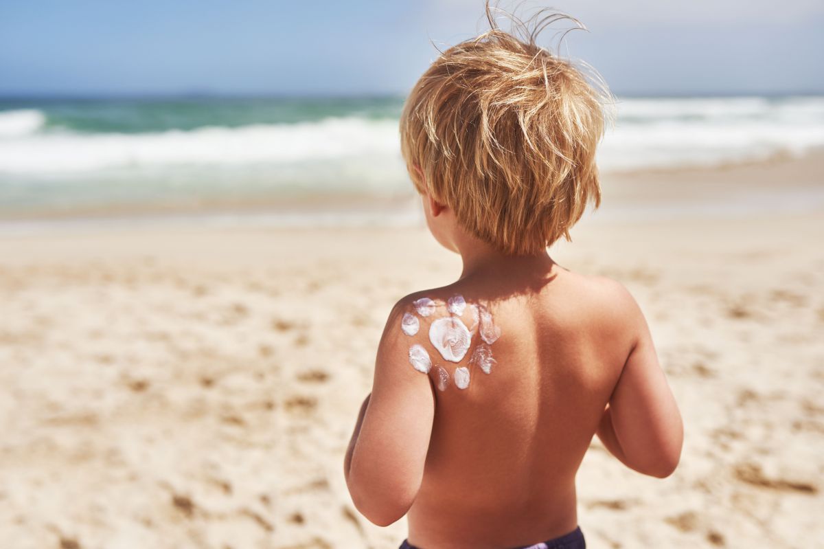 Ein Kind steht an einem Strand in der Sonne. Auf seine Schulter ist mit Sonnencreme eine Sonne aufgemalt.
