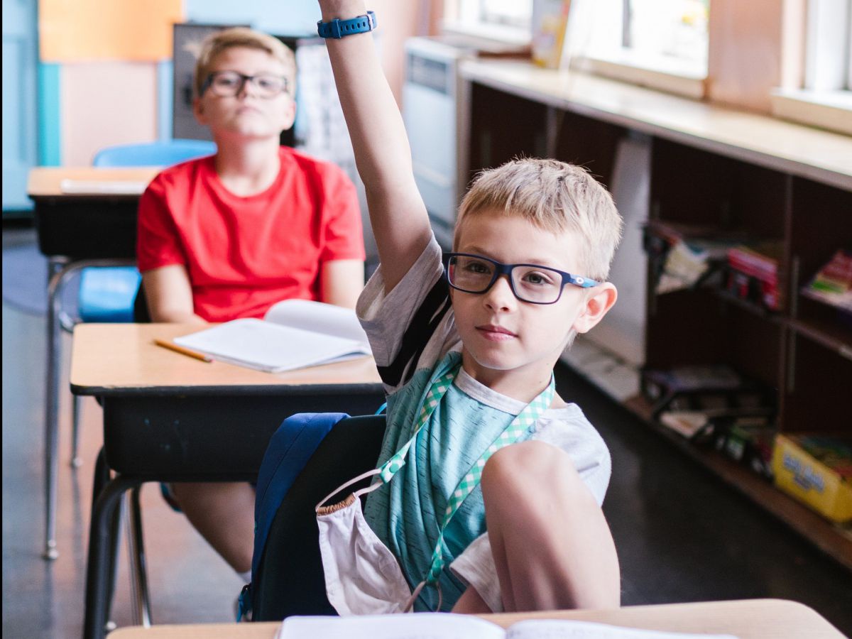 Zwei Kinder mit Brillen in einem Klassenzimmer. Beide blicken nach vorne, eines hebt die Hand.