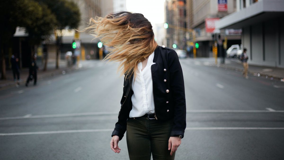 Eine Frau in schwarzen Jeans, weißer Bluse und schwarzer Jacke steht mitten auf der leeren Fahrbahn einer Straße in einer Großstadt. Eine starke Windböe bläst ihr das blonde lange Haar vors Gesicht.