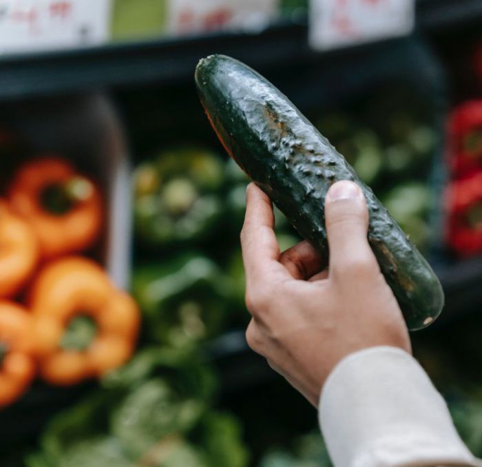 Vor dem Hintergrund einer Gemüsetheke hält eine männliche Hand eine Feldgurke. Am Arm hängt ein blauer Kunststoffeinkaufskorb.