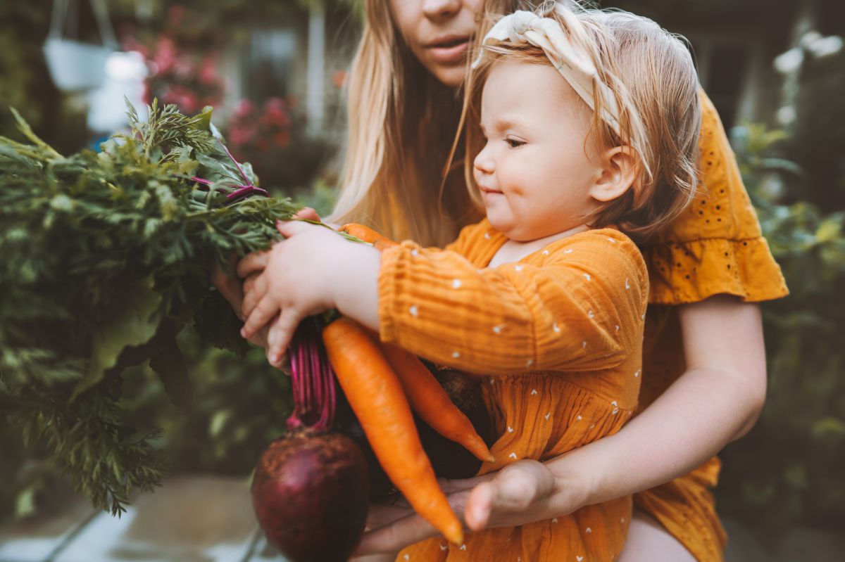 Ein Kind, gehalten von den Armen der Mutter, hält einen Bund Karotten und Zwiebel in den Händen.