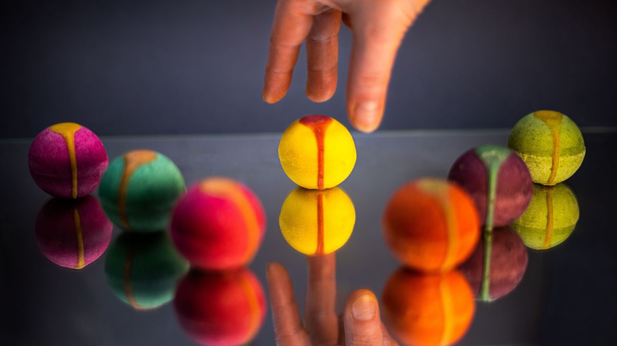 Auf einer spiegelnden Oberfläche sind sieben bunte Kugeln symmetrisch aufgelegt. Eine Hand greift die mittlere Kugel.