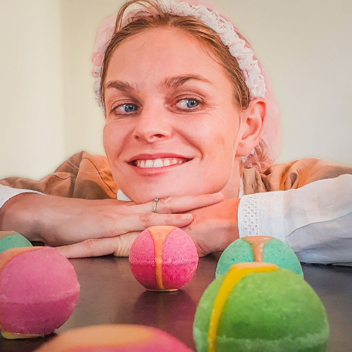 Eine Frau mit blonden langen Haaren und blauen Augen stützt ihr Gesicht auf den Händen ab. Auf dem Tisch vor ihr liegen bunte Badekugeln.