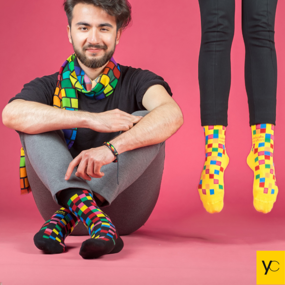 Ein junger Mann mit Bart sitzt auf dem Boden, er trägt auffällig bunte Socken und einen ebensolchen Schal. Neben ihm sieht man zwei Beine ins Bild hängen, an den Füßen ebenfalls bunte Socken. Der Bildhintergrund ist in pink gehalten.