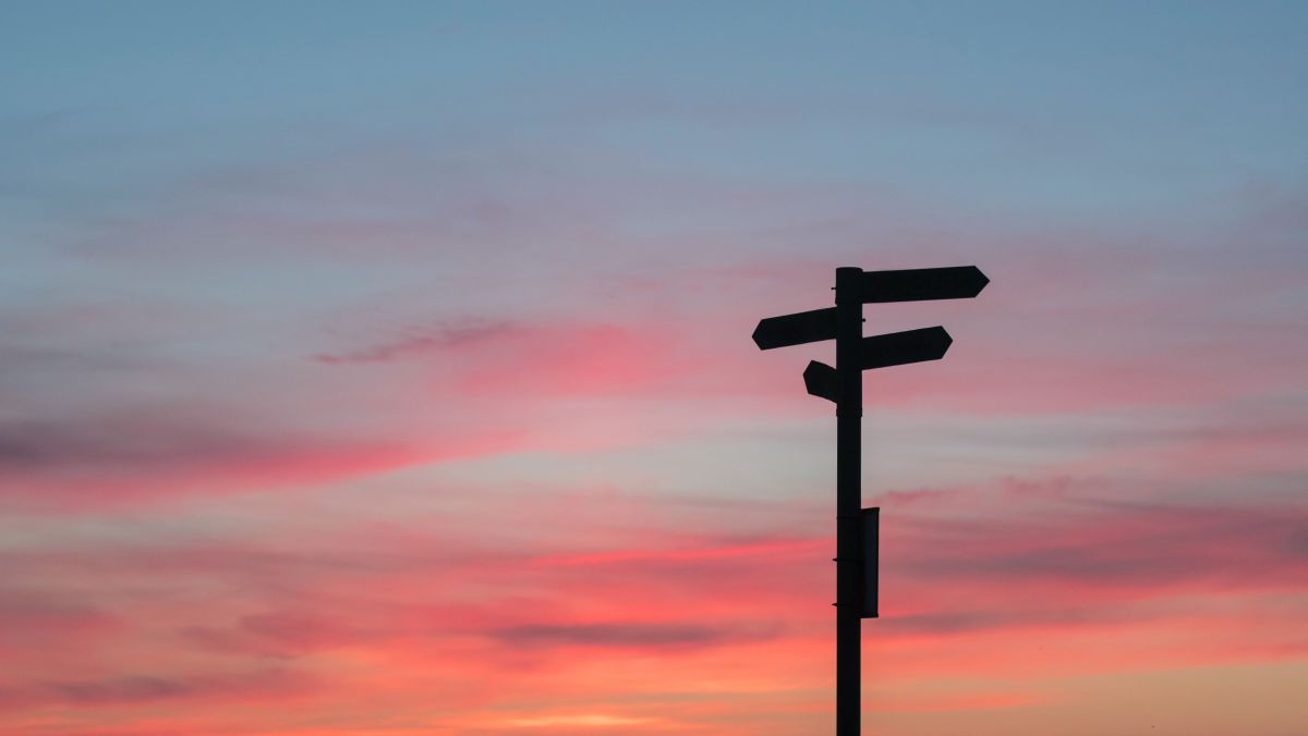 Ein Wegweiser zeichnet sich schwarz vor den Farbverläufen des Sonnenuntergangs ab.