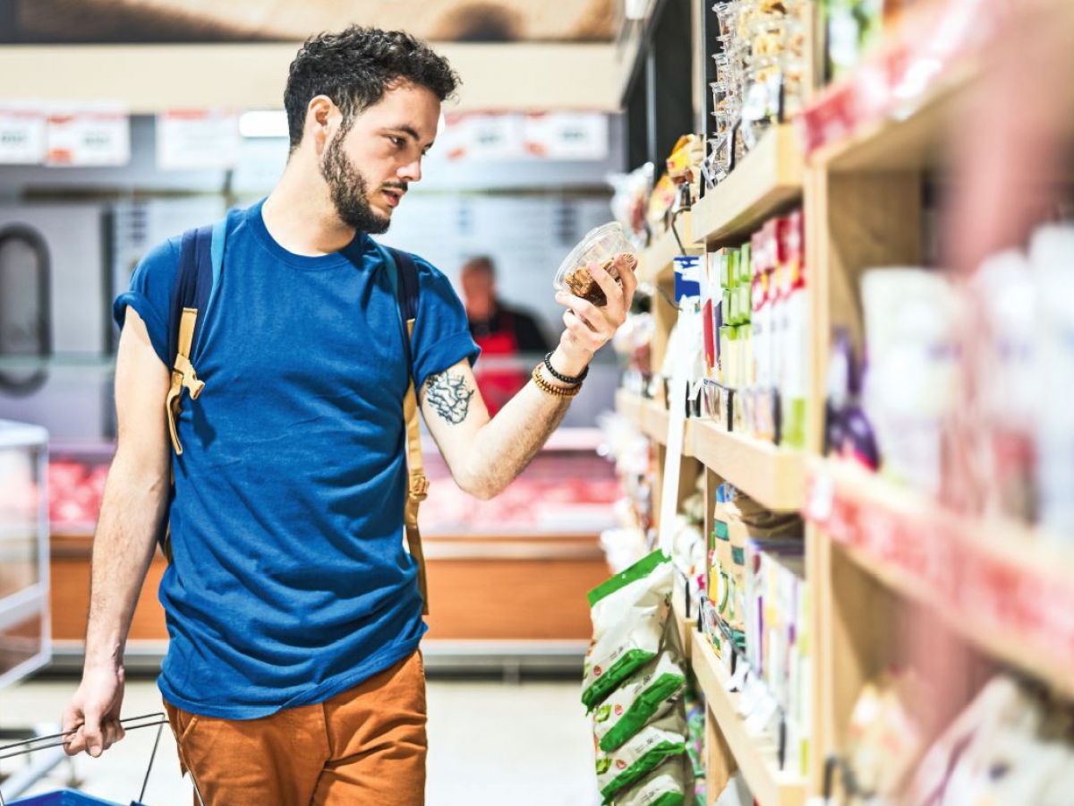 Ein Kunde in einem Supermarkt steht vor dem Regal und begutachtet die Aufschrift der Verpackung eines Lebensmittels.
