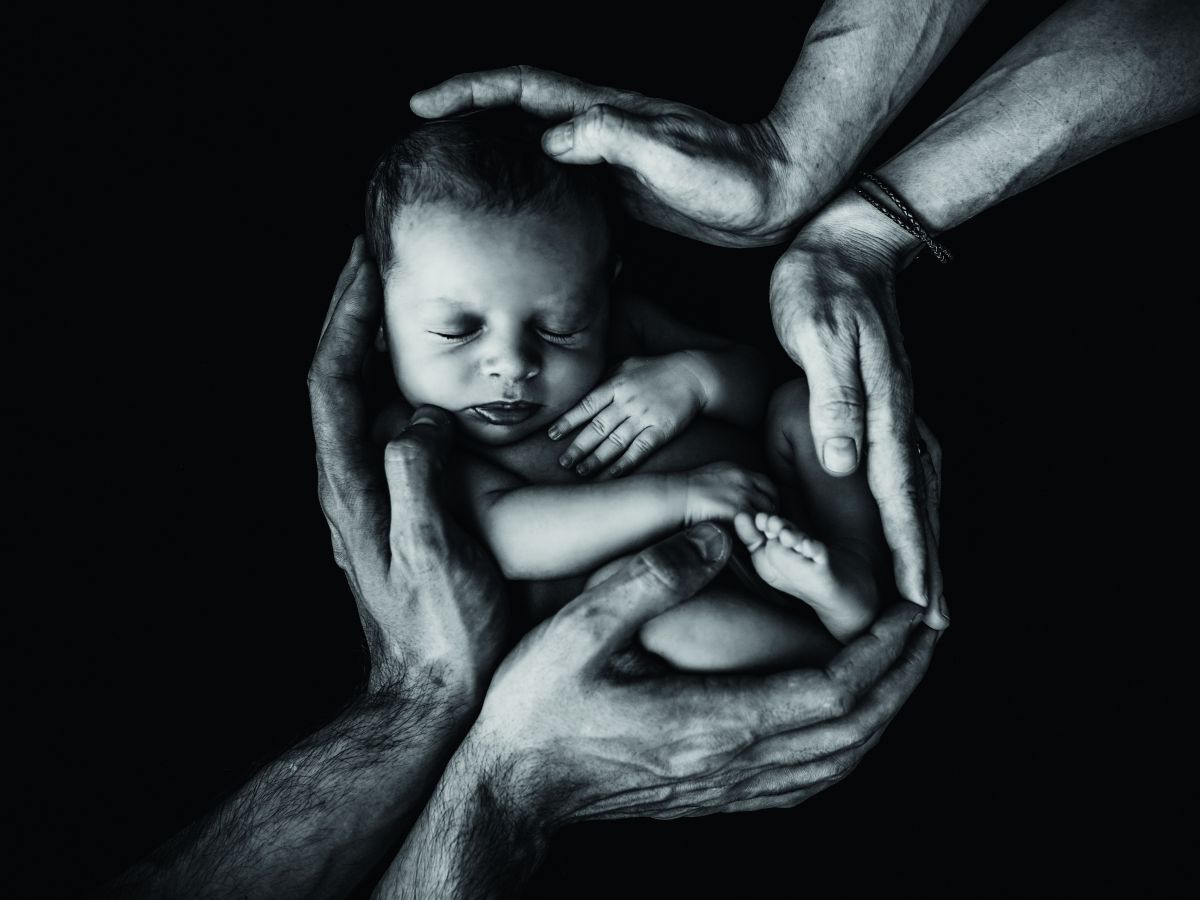 Foto in kontrastreichem Schwarz-weiß: Ein Neugeborenes, das von den Händen zweier Männer schützend umfangen wird.