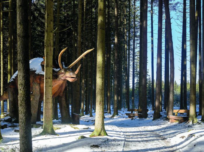 Eine lebensgroße Dinosaurierfigur steht zwischen Baumstämmen am winterlich schneebedeckten Erlebnisweg Moorwald. 