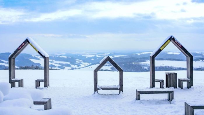 Hölzerne Sitzgelegenheiten in Form stilisierter Häuser auf dem schneebedecktem Gipfel eines Hügels. Der Blick hinunter ins Tal fällt auf eine winterliche Landschaft mit Wiesen und Wäldern.