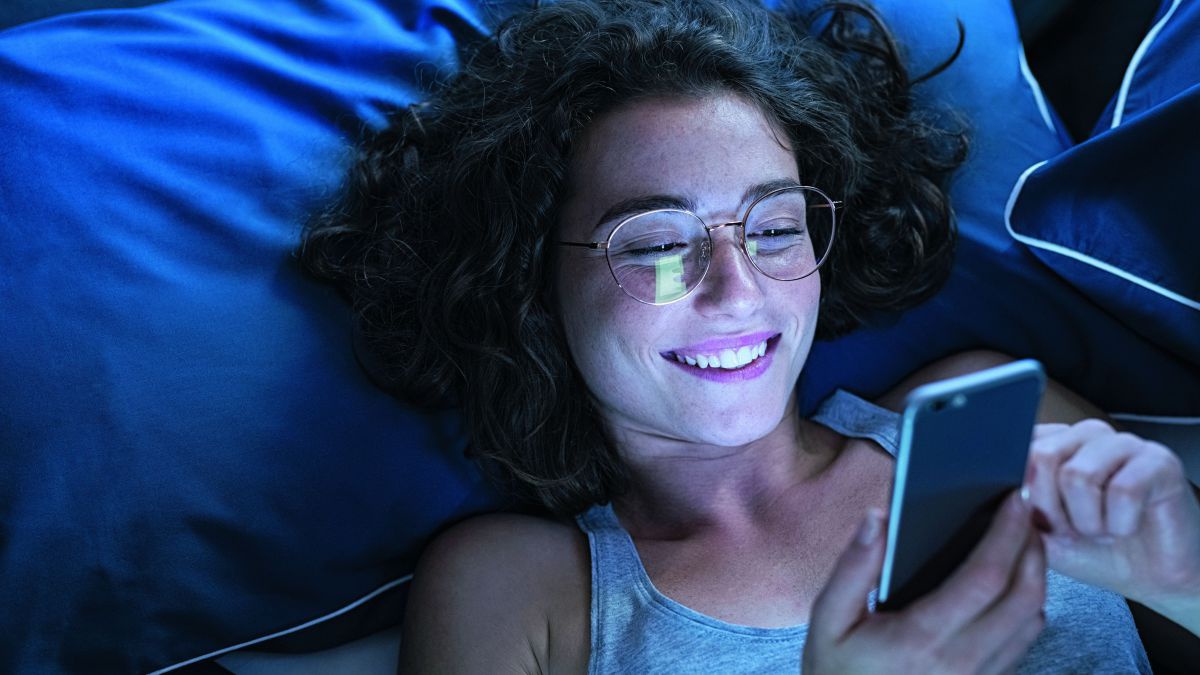 Eine junge Frau mit Brille liegt im Bett und schaut lachend etwas auf ihrem Smartphone an. Die Bettwäsche ist dunkelblau, das gesamte Bild ist in blaues Licht getaucht.