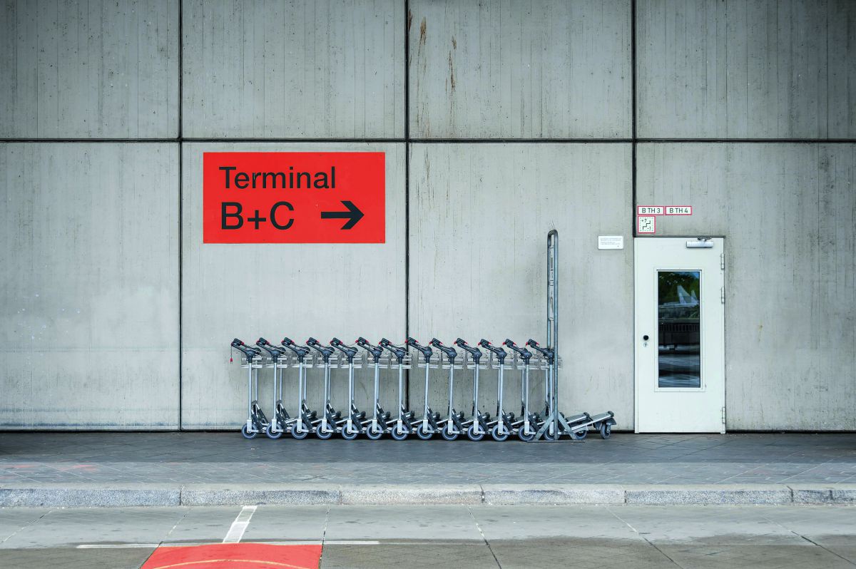 Gepäcktrolleys vor dem Flughafengebäude. Ein oranges Schild an der Betonwand trägt Aufschrift mit Richtungspfeil:  Terminal B+C.