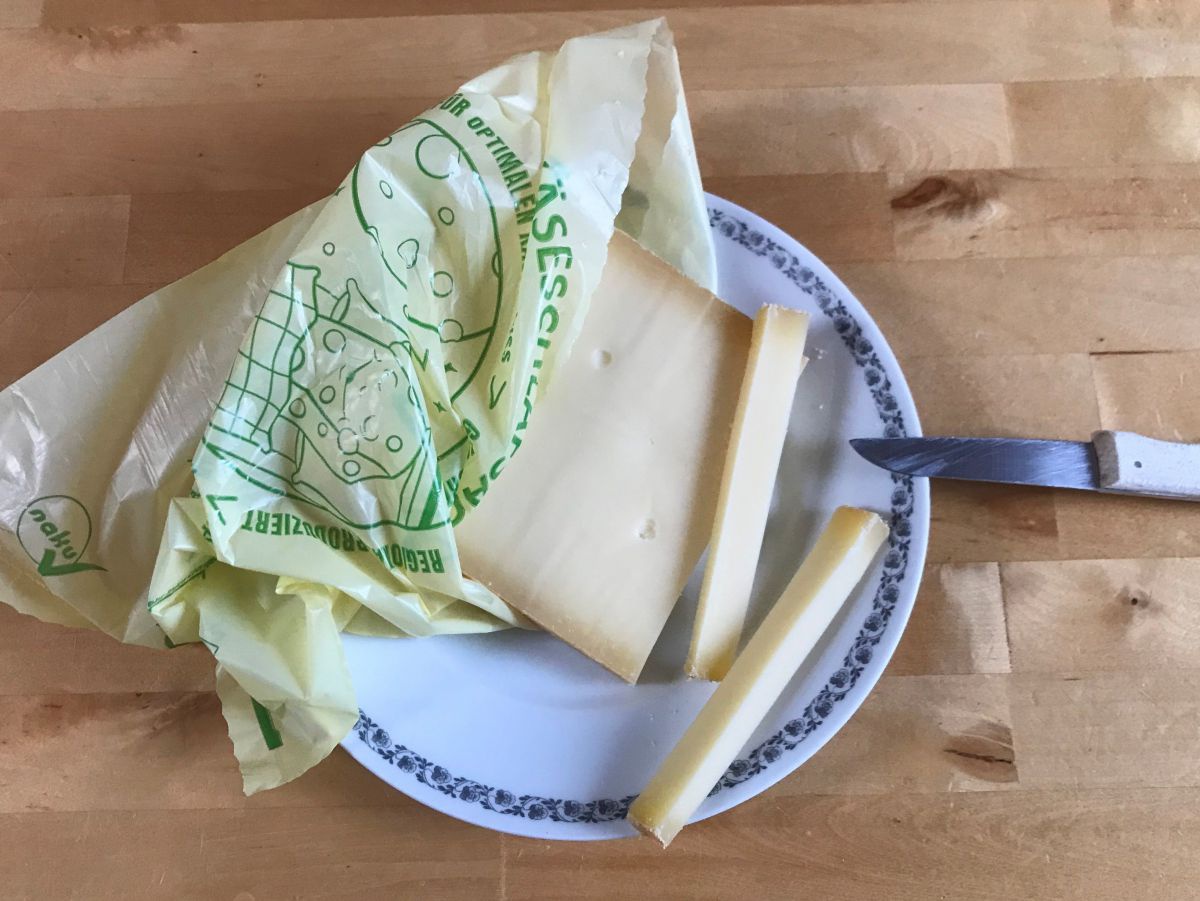 Käse auf einem Teller, ragt zur Hälfte aus dem Naturkunststoffbeutel, es wurden zwei Schnitten abgetrennt, Messer liegt am Teller.