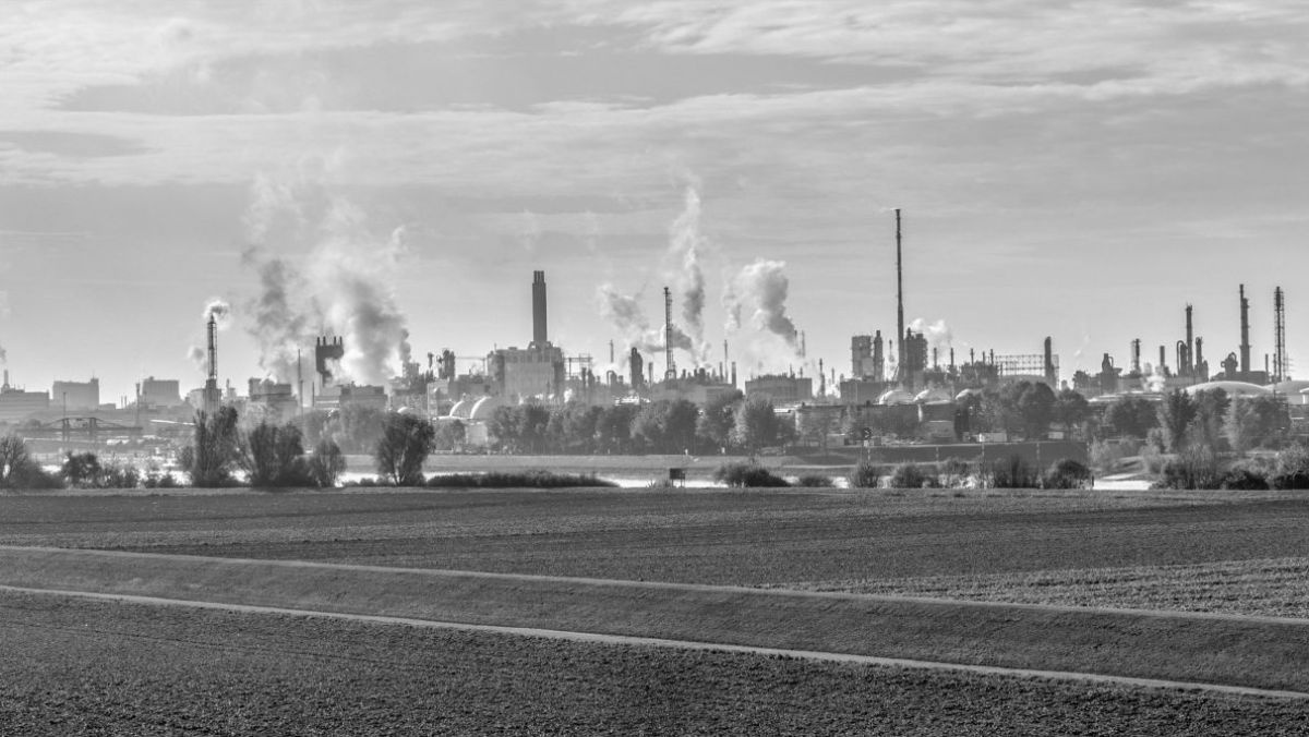 Ein schwarzweiß Bild einer Industriezone. Im Hintergrund sind Fabriken und Schornsteine zu sehen, im Vordergrund liegen Felder.