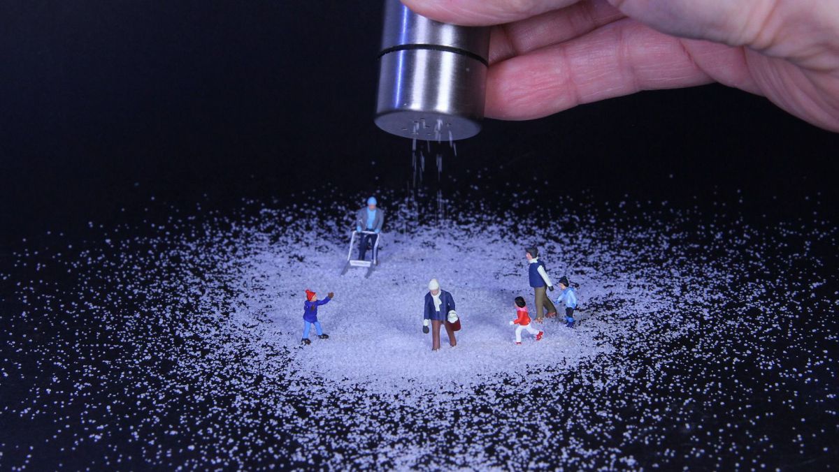 Auf einem schwarzen Untergrund stehen Miniaturfiguren in Winterkleidung. Eine Hand salzt sie mit einem Salzstreuer aus Metall.