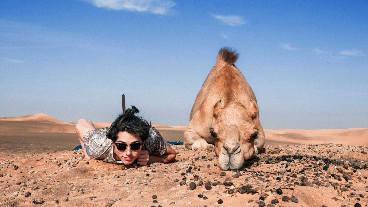In der Wüste liegen eine Frau und ein Kamel nebeneinander auf dem Bauch am Boden und blicken in die ebenfalls am Boden stehende Kamera.