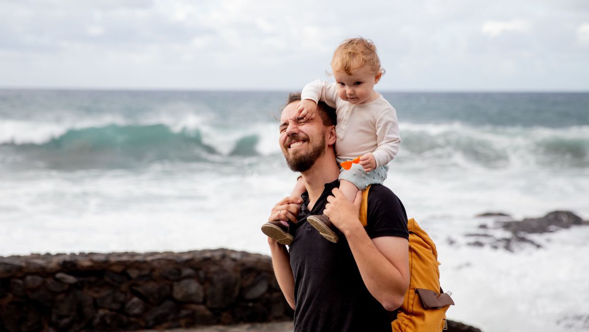 Ein junger Vater trägr sein etwa einjähriges Kind auf den Schultern, das ihm mit den Fingern unbewusst am Auge kneift. Im Hintergrund ein felsiger Strand und Meeresbrandung.