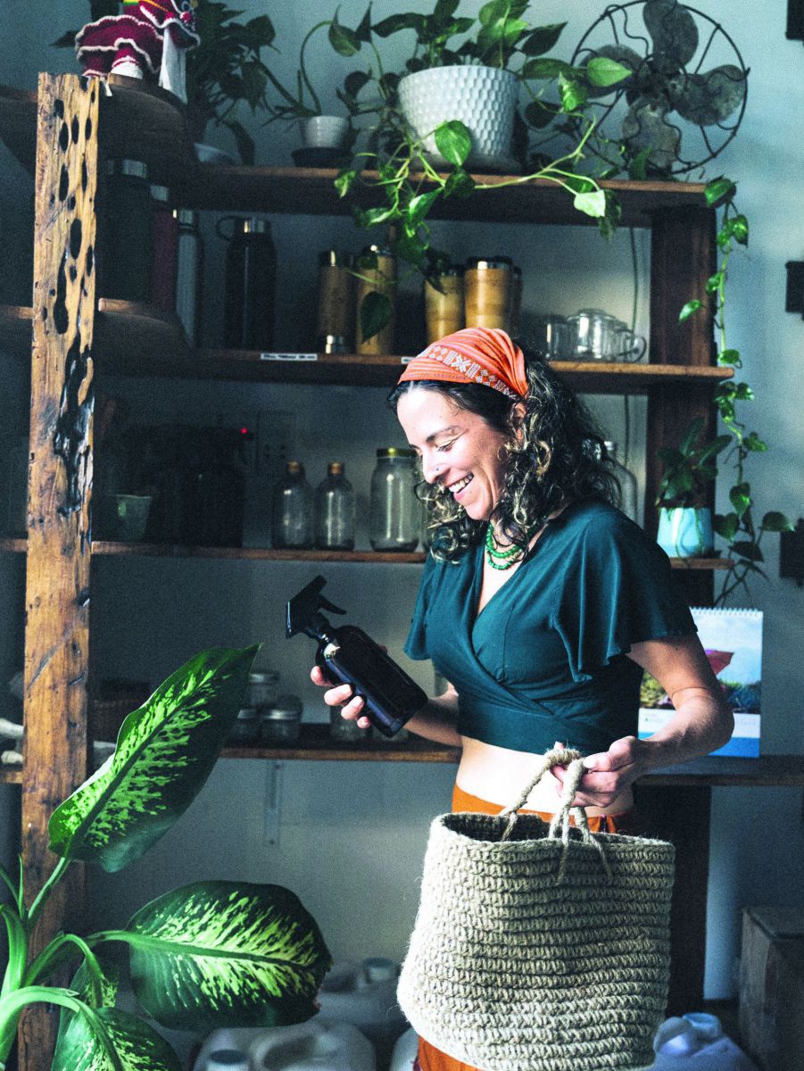 Eine Frau steht vor einem Regal in einem Verkaufsraum und hält eine Sprühflasche in der einen und einen Einkaufskorb in der anderen Hand.