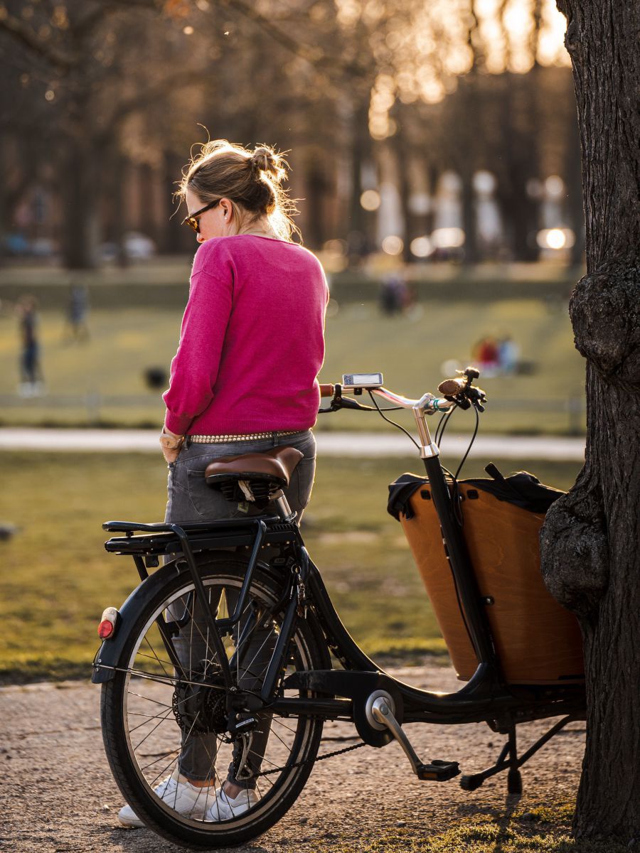 In einem Park mit alten Bäumen steht eine Frau mit pinkem Pullover mit dem Rücken zum Betrachter an ihr Lastenfahrrad gelehnt.