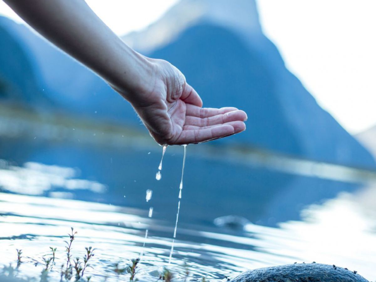 Am Ufer eines Gebirgssees, jemand schöpft Wasser mit der Hand, ein dünnes Rinnsal tropft von den Fingern zurück in den See und auf einen runden großen Stein.
