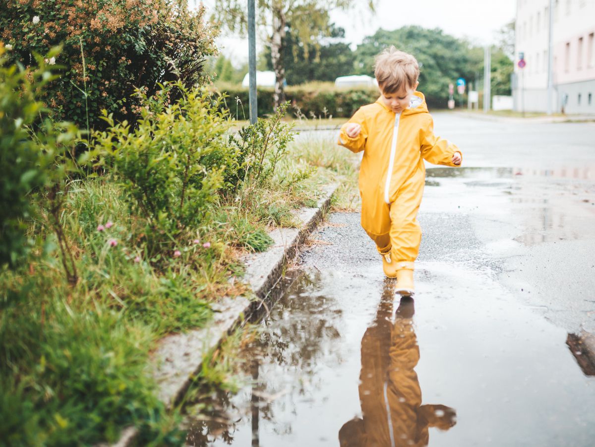 Es hat geregnet. Ein Kleinkind in einem gelben Regenoverall und Gummistiefel läuft durch eine große, spiegelnde Wasserlacke am Straßenrand.