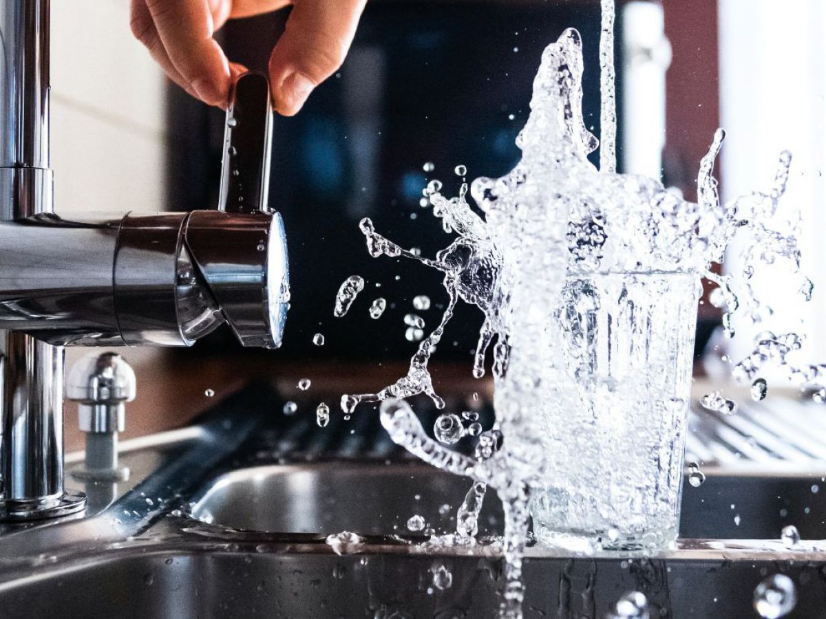 Ein Glas wird unter einem Wasserhahn in einer Küche mit hohem Druck mit Leitungswasser befüllt, so dass das Wasser übersprudelt.