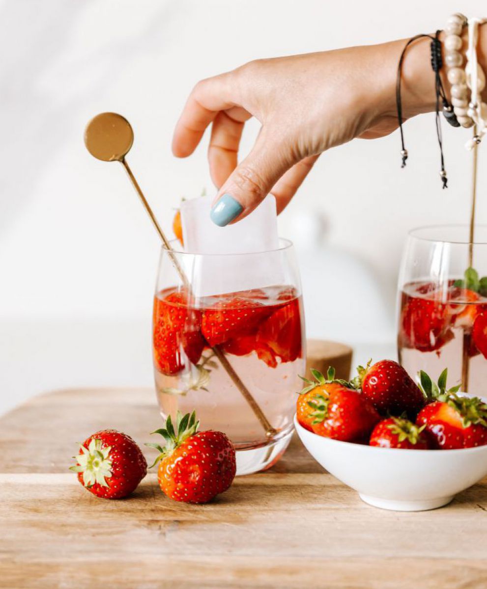 Erdbeerwasser: In Trinkgläsern schwimmen halbe Erdbeeren in Wasser, eine Hand mti hellblau lackierten Fingernägeln und Armbändern lässt gerade einen großen Eiswürfel ins Glas fallen.