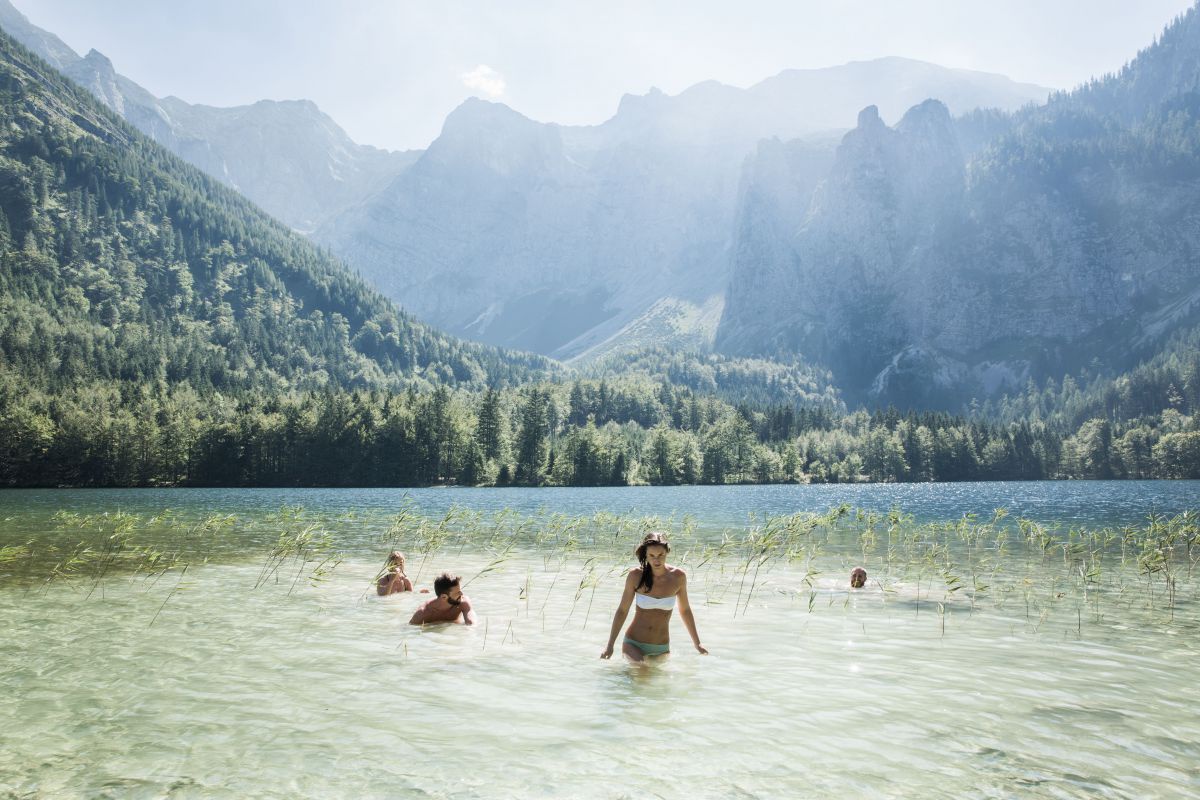 Badende im seichten Uferbereich des Hinteren Langbathsees, im Hintergrund das Bergpanorama des Salzkammergutes, die Sonnenstrahlen fallen steil ein und lassen das Wasser und die Luft glitzern.