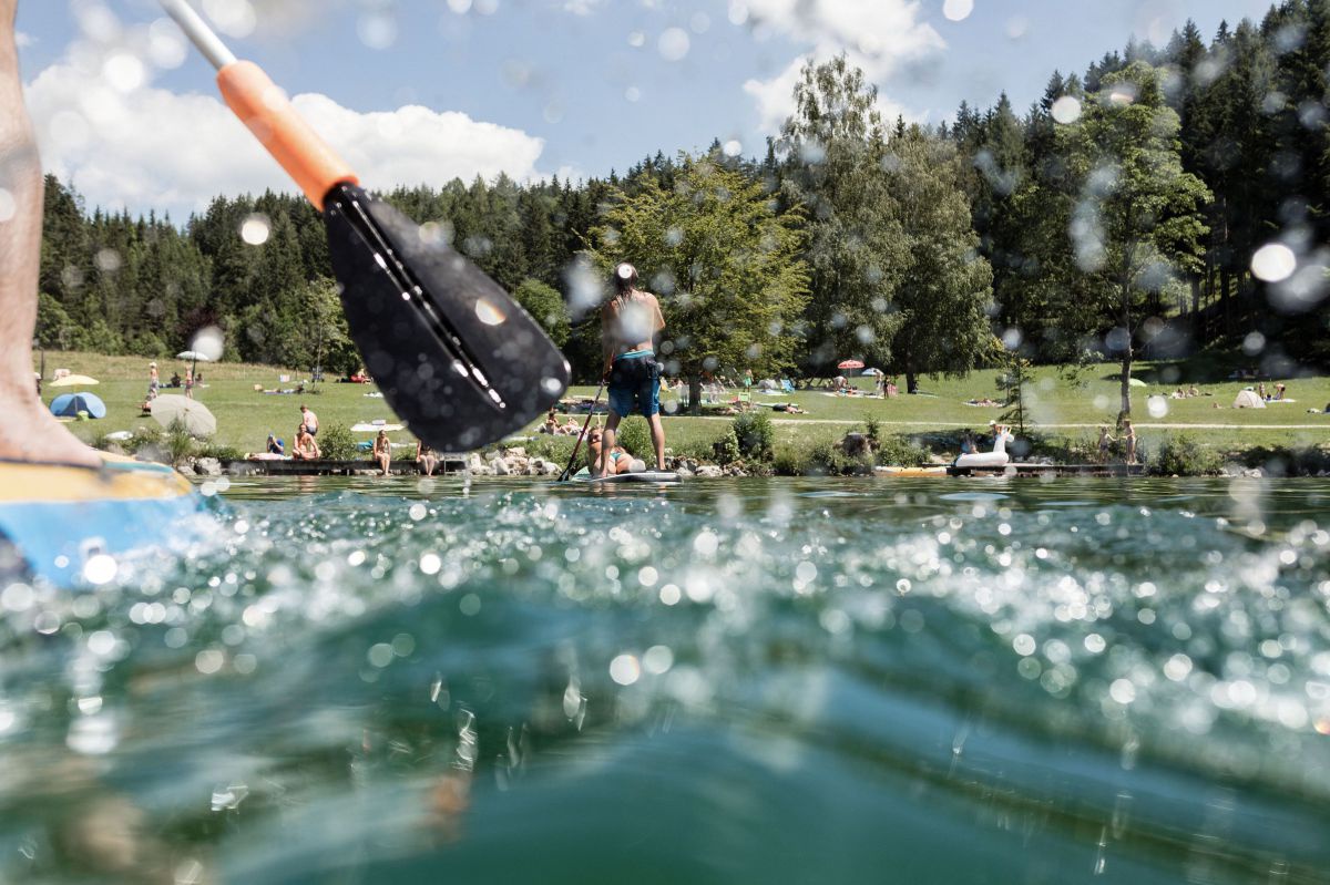 Fotoaufnahme knapp über der Wasseroberfläche ganz nah am Paddel eines Stand-up-Paddlers, das Wasser spritzt auf die Linse, im Hintergrund sieht man das bunte Treiben von Wassersportlern und die bewaldete Liegewiese am Seeufer.