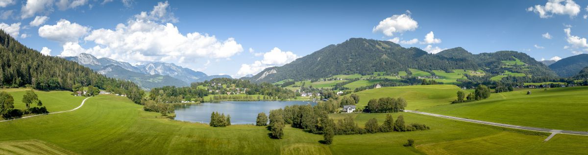 Der Putterersee liegt eingebettet in die malerische Landschaft aus grünen Wiesen und Wäldern, im Hintergrund Berge und ein strahlend blauer Himmel mit weißen Wolken.