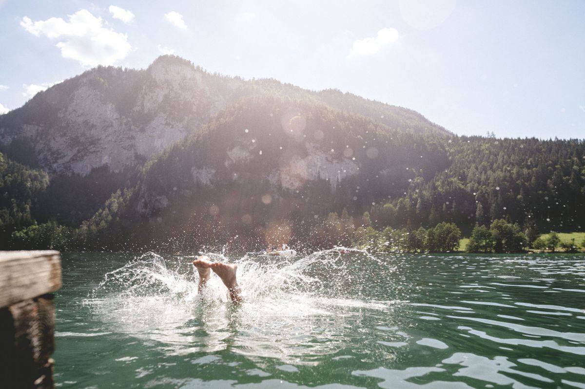 In einen See springt gerade jemand so von einem Holzsteg, dass nur die Wasserspritzer in der Sonne und die Füße erkennbar sind.
