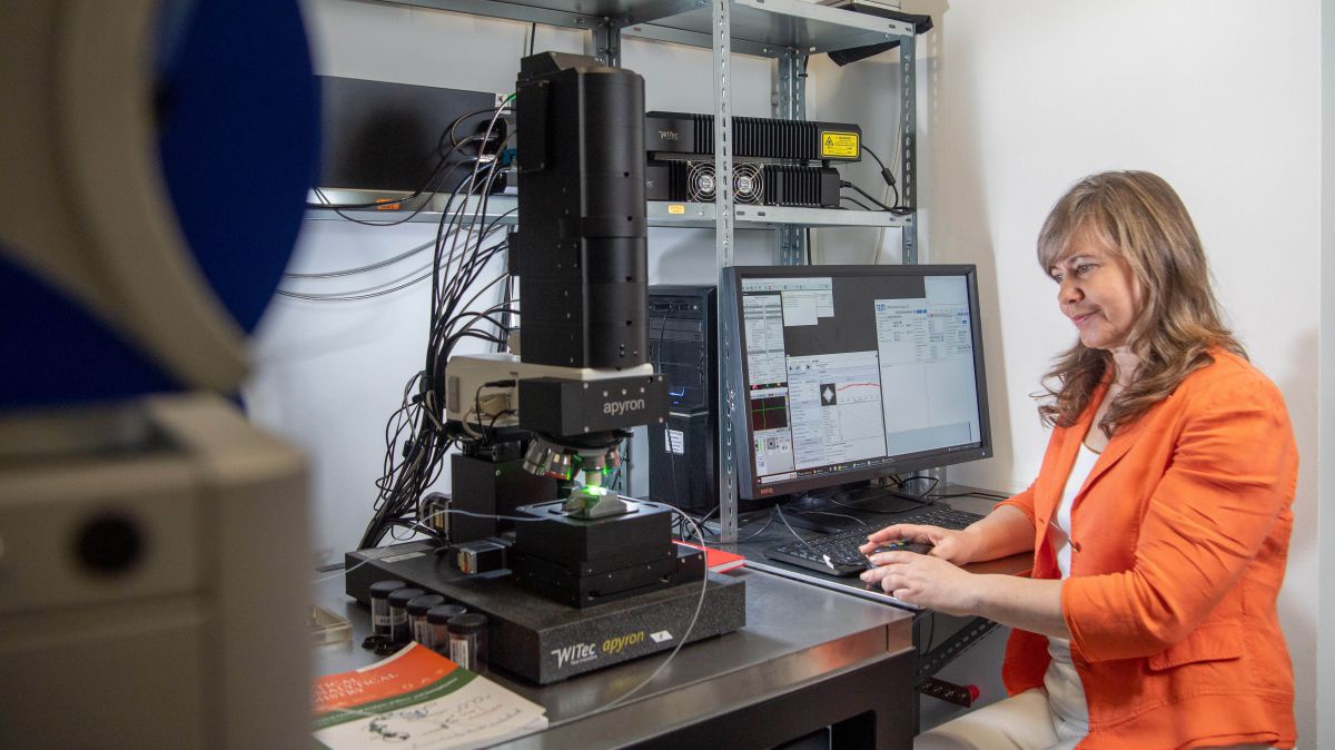 Eine Frau mit orangenem Blazer und langen blonden Haaren arbeitet an einem Mikroskop-Computer.