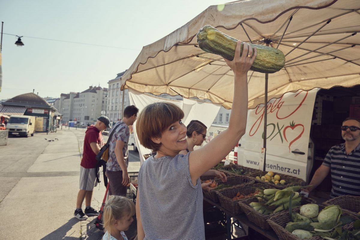 Eine Frau steht vor einem Marktstand und hält eine große Zuccine hoch.
