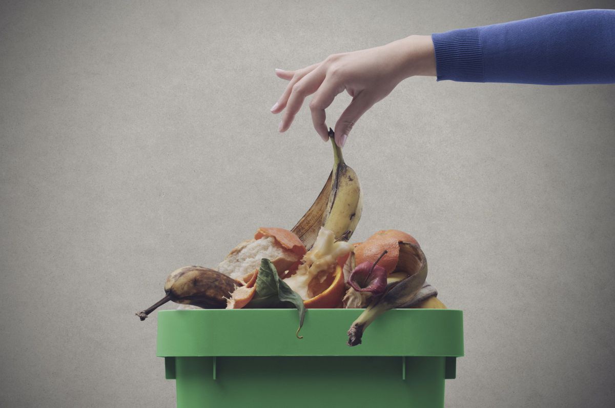 Eine Mülltonne mit überquellendem Biomüll, eine Hand hebt mit spitzen Fingern eine Bananenschale hoch.