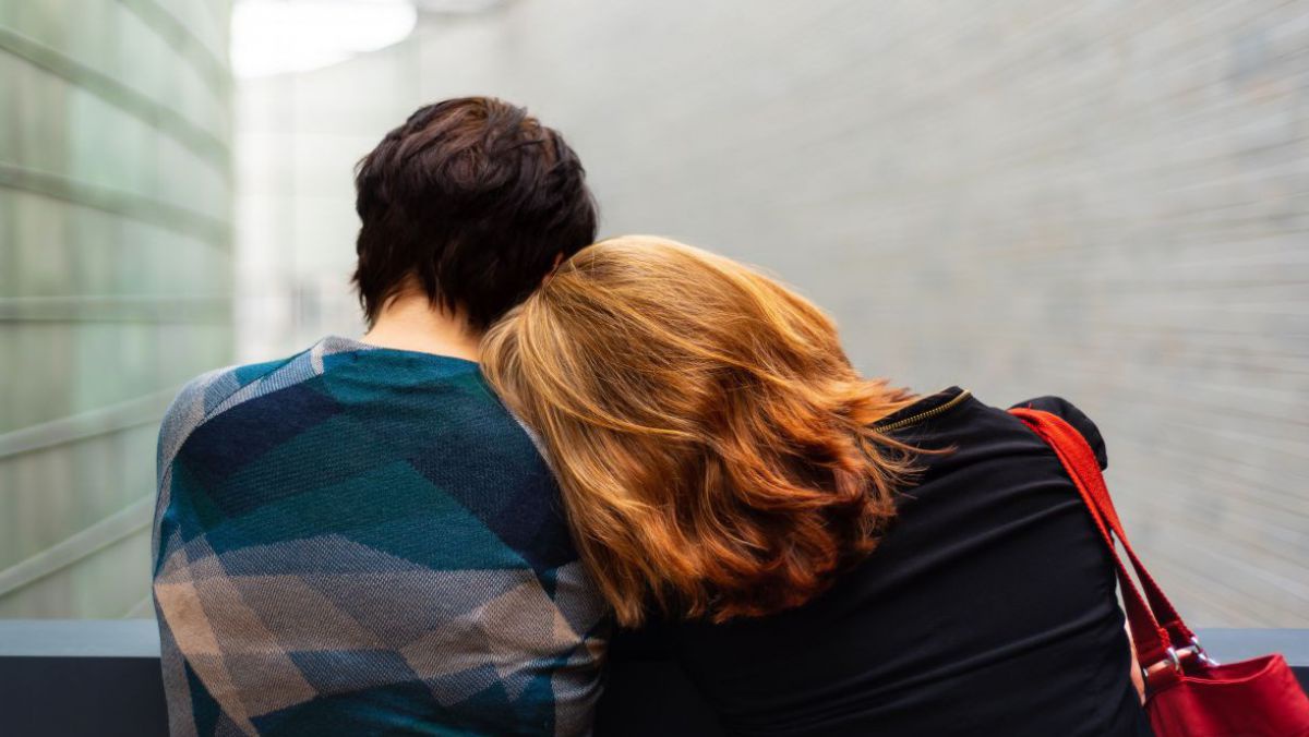 Wir sehen zwei Menschen nebeneinander sitzen, sie drehen der Kamera den Rücken zu. Die linke Person trägt ihr dunkles Haar kurz geschnitten und hat einen blaugemusterten Pullover an. Die rechte Person trägt ein dunkelblaues Oberteil und eine rote Handtasc