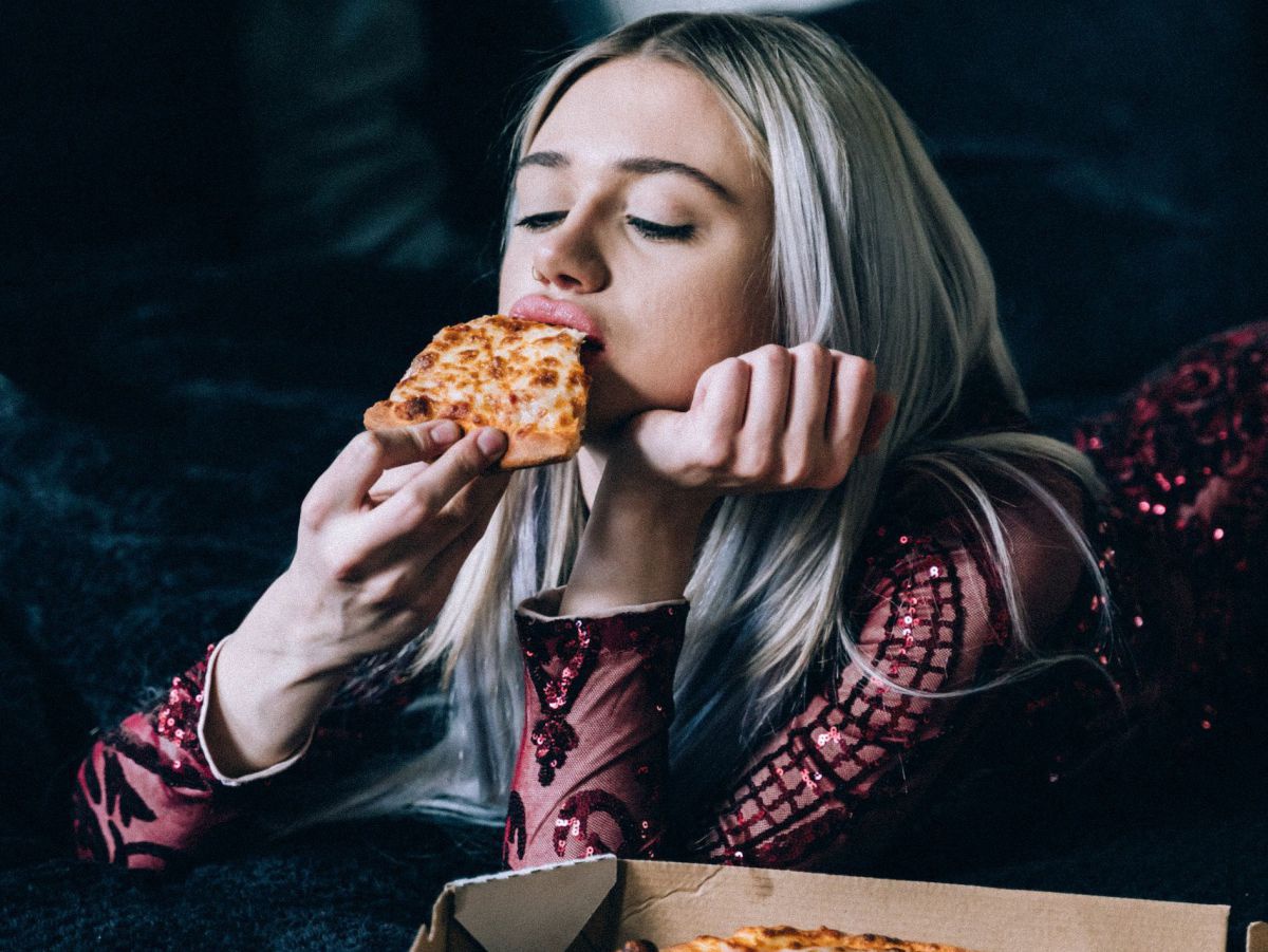 Eine junge Frau liegt bäuchlings auf einem Bett und beißt in in Stück Käsepizza. Sie ist gekleidet und geschminkt wie für einen feierlichen Anlass. Ihr Gesichtsausdruck wirkt bedrückt. Der Pizzakarton liegt neben ihr auf dem schwarzen Bettüberwurf.