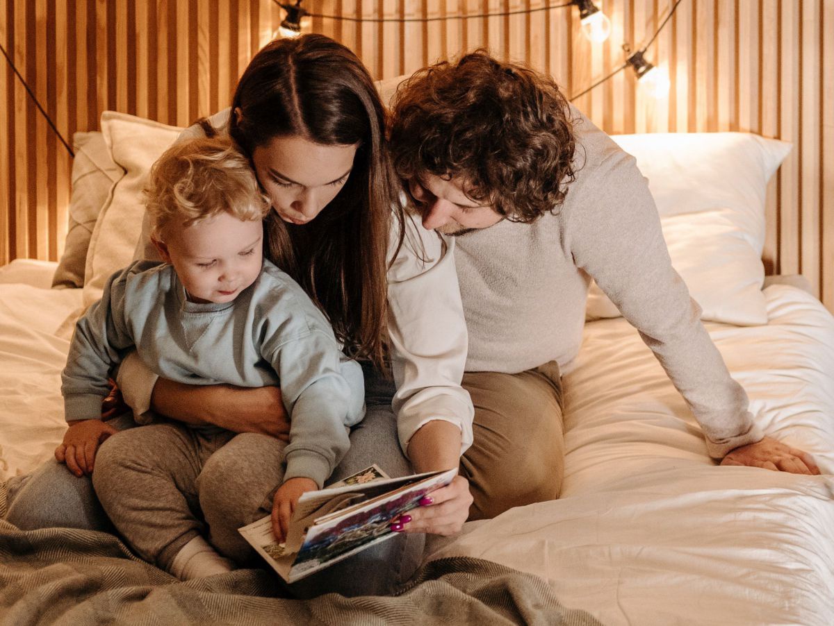Ein Paar mit Kleinkind kuschelt zusammen auf dem Familienbett und schmökert in einem Bilderbuch.