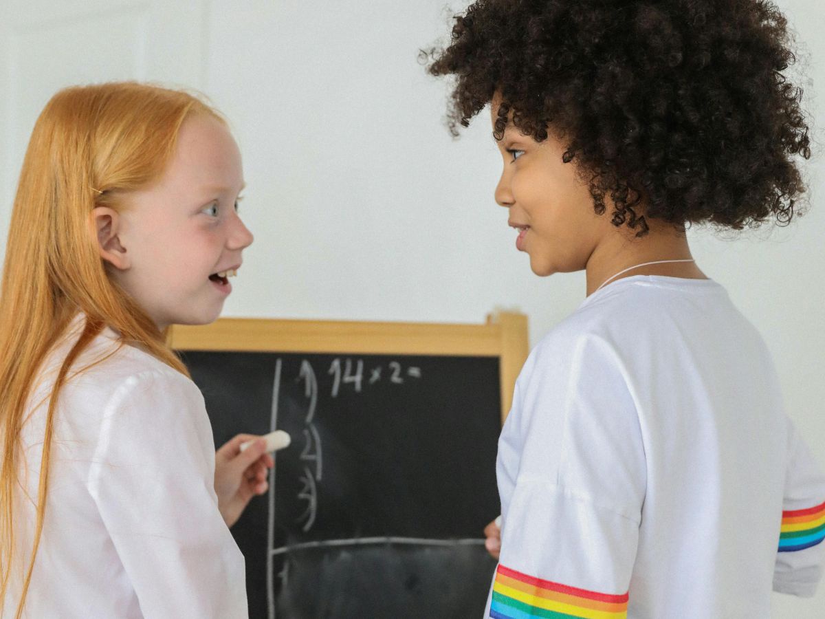 Zwei Mädchen im Grundschulalter schreiben mit Kreide Multiplikationen an eine Tafel, sie sehen einander dabei lachend kommunizierend an.